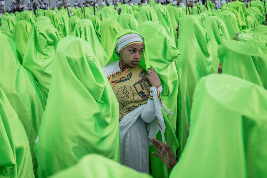 A person turns toward the camera, standing among a crowd of people wearing long green hooded garments.
