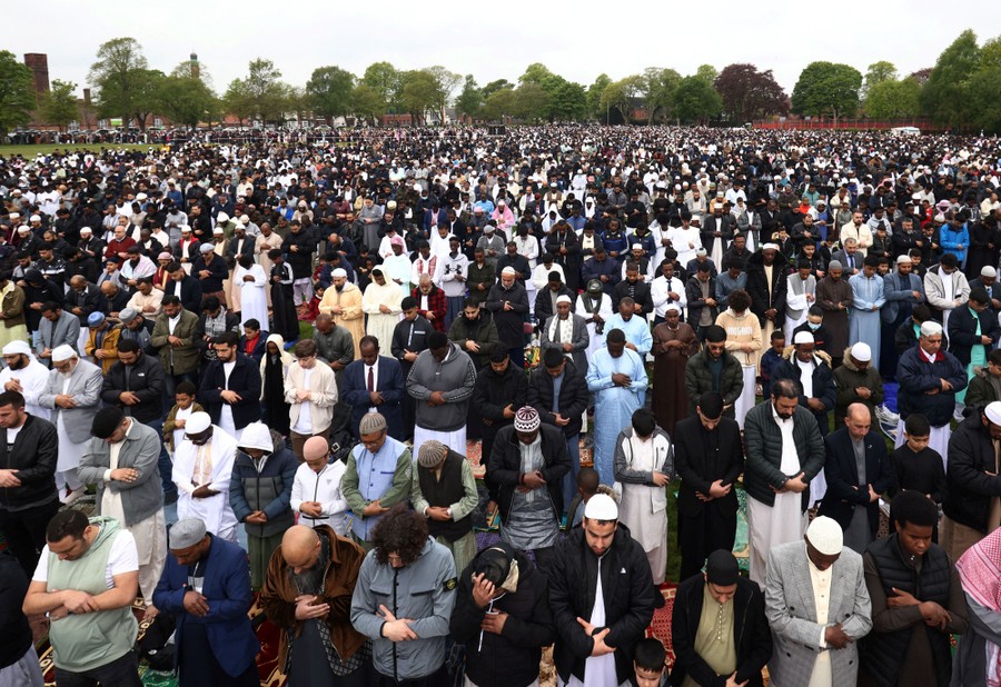 A large crowd of men stands in prayer in a park.