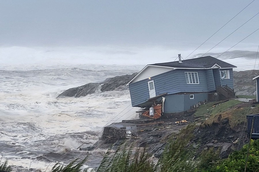 A damaged house leans sharply toward a storm-driven surf.