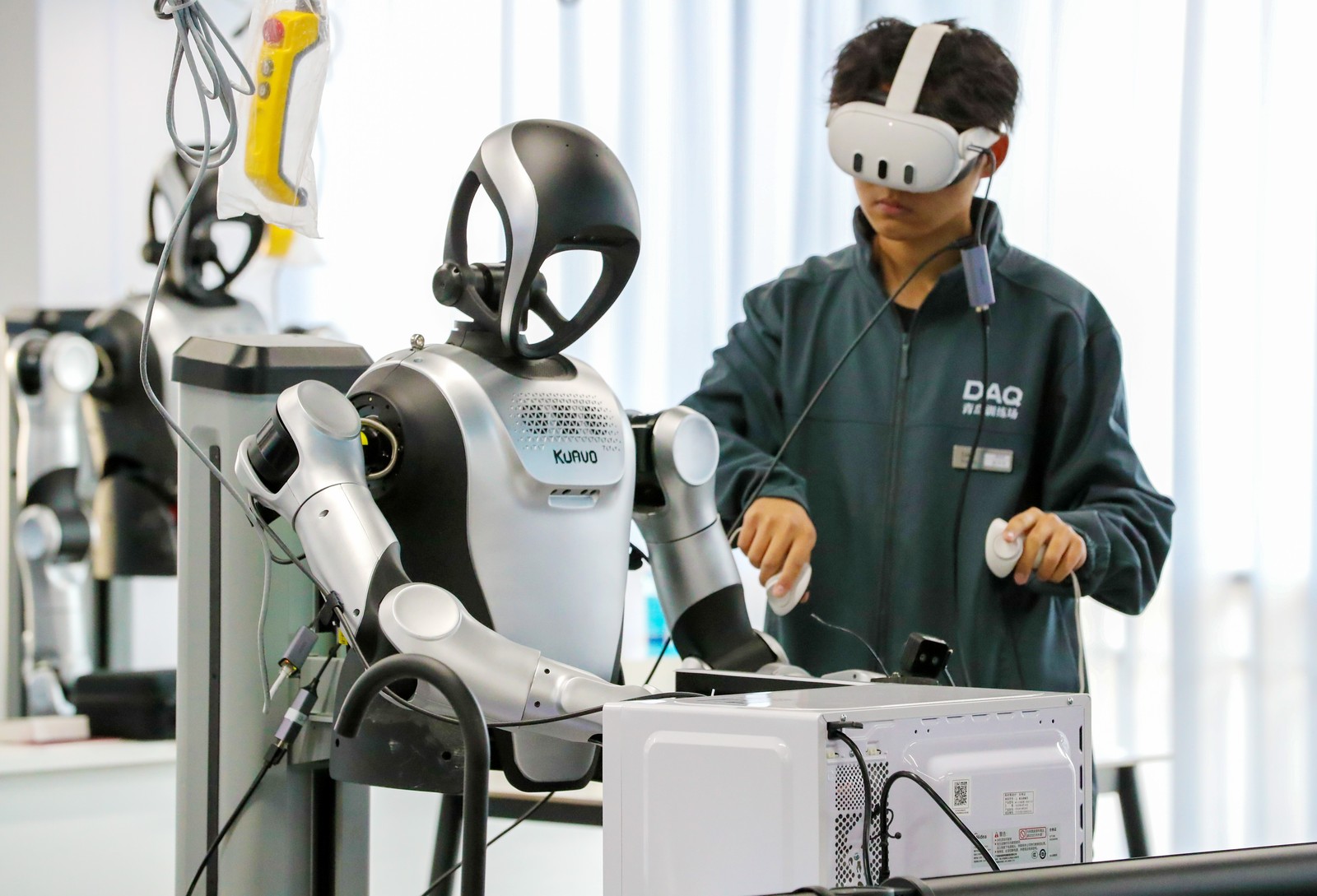 A person wearing VR goggles stands beside a humanoid robot, training it to clean a microwave oven.