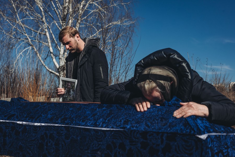 A person leans over a coffin, as another person stands nearby, holding a small portrait.