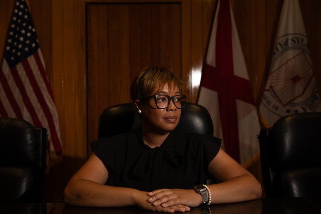 A woman sits in a black chair leaning on a desk wearing a black shirt