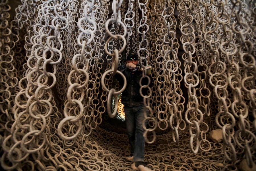 A worker stands inside a large-diameter pipe with dozens of large chains hanging from the ceiling.