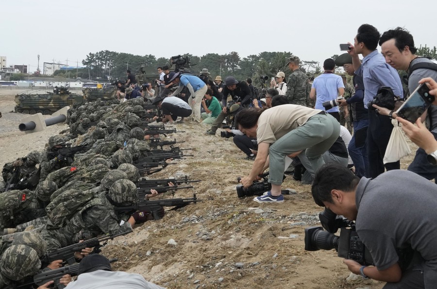 Many members of the media crouch and film close to a line of soldiers lying along a berm on a beach.