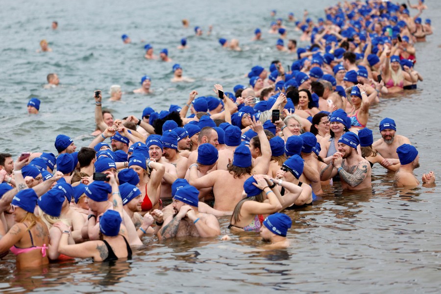 Hundreds of people, most wearing blue bathing caps and bathing suits, stand or swim in chest-deep water.