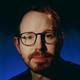 Ari Aster wearing glasses against a blue background