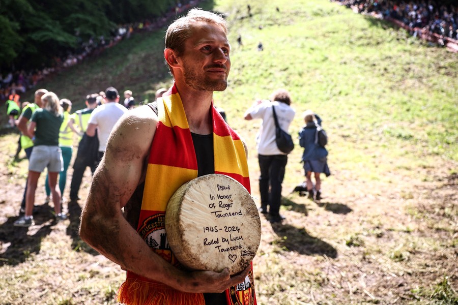 A muddy but cheerful man poses at the bottom of a steep hill, holding his prize, a round of cheese. Written on the round are the words "Rolled In Honor Of Roger Townsend, 1945–2024. Rolled by Lucy Townsend."