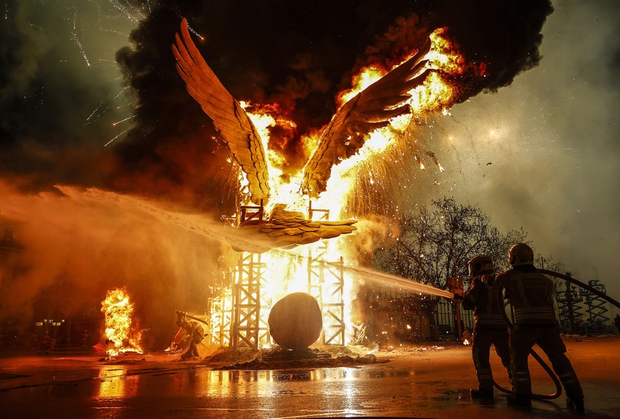 Two firefighters hold a hose, spraying water near a large burning sculpture.