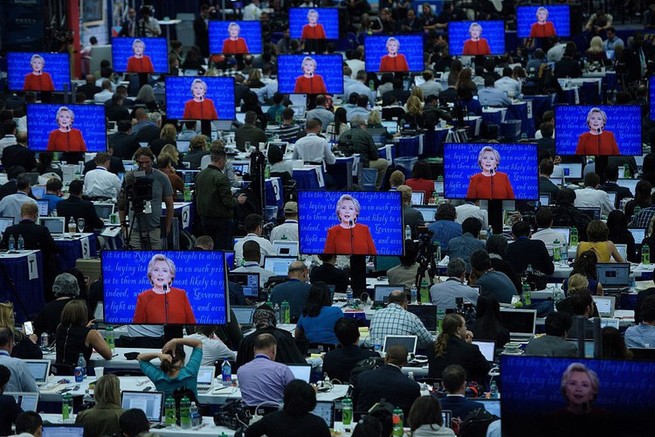 a crowd of people working in front of many monitors broadcasting Hillary Clinton
