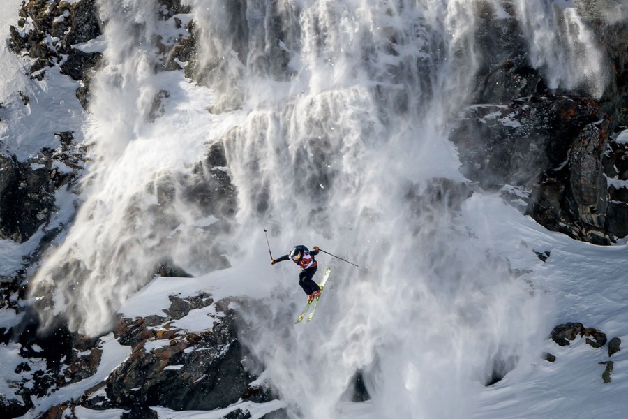 A skier makes an enormous jump along a rocky mountainside with snow cascading down the rocks behind him.