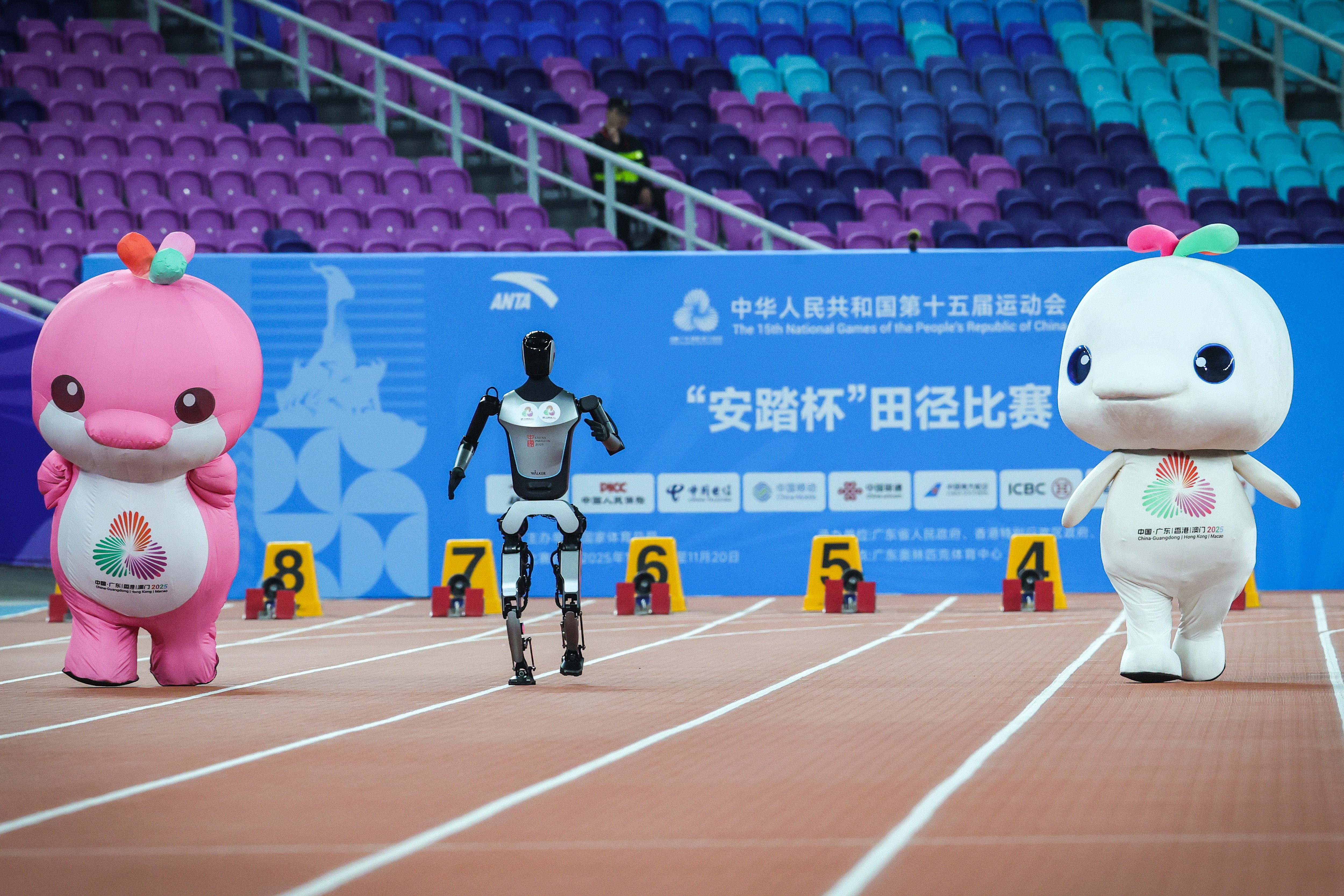 Two people in mascot costumes run on a track, racing against a humanoid robot.
