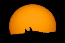 Two people sit on a hillside, silhouetted by the setting sun.