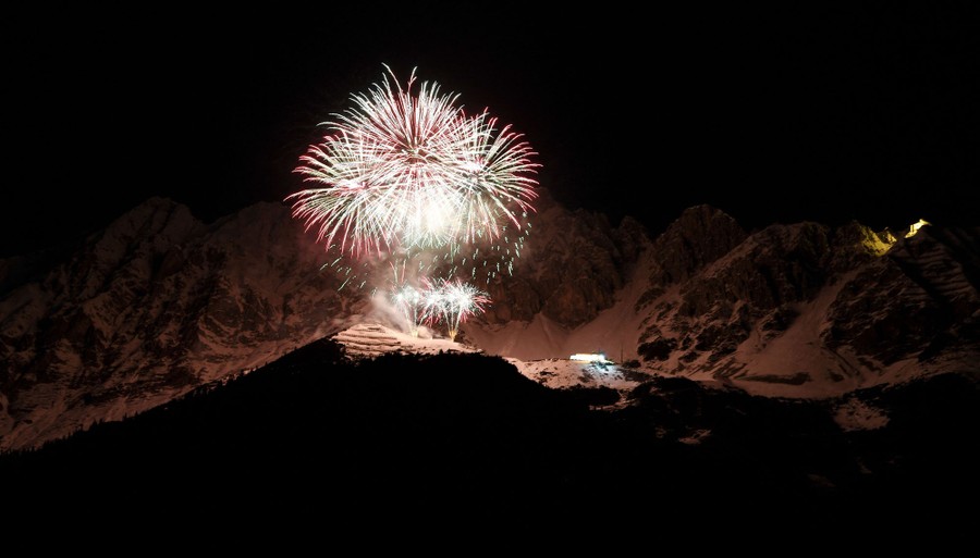 Fireworks explode above a mountaintop, illuminating more mountains in the background.