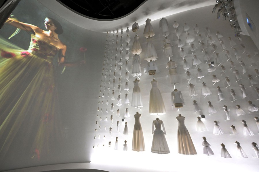 An exhibition space on a curved wall, featuring a large projected image of a model in a dress on the left side and many small and large dresses mounted on the wall on the right side