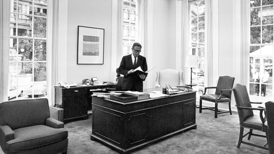 Henry Kissinger standing at a desk in an office with high windows in 1970