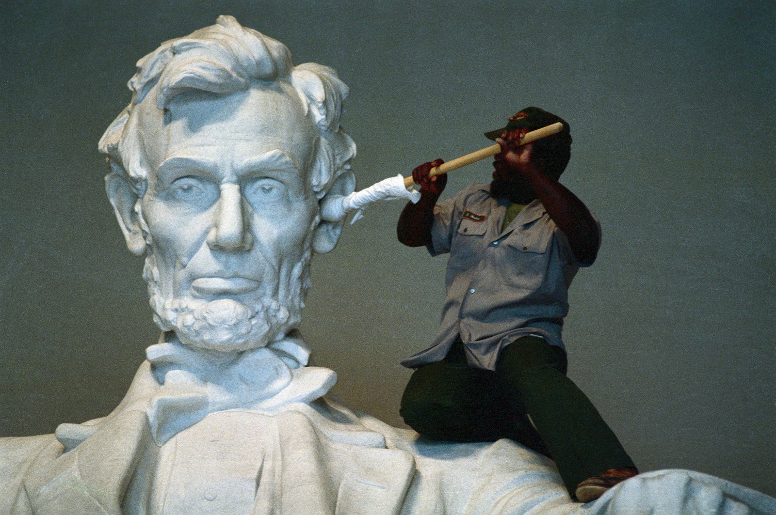 A person uses a cloth-wrapped pole to clean the ear of a large statue of Abraham Lincoln.