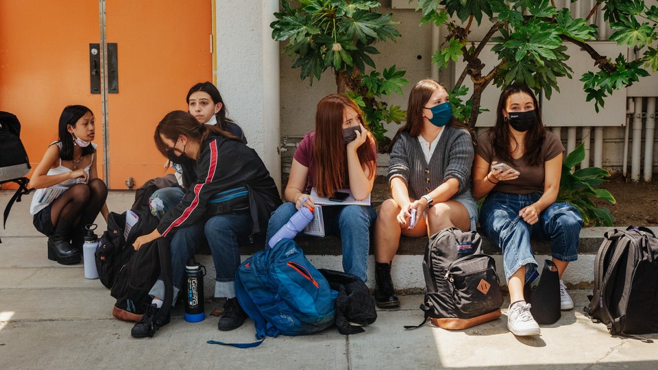 High-school students wearing masks sit together outside a school building.