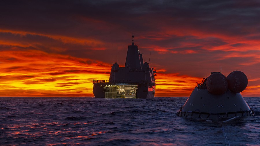 A space capsule floats in the ocean, near a U.S. Navy vessel, at sunset.