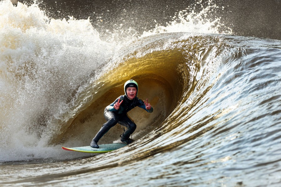A young surfer holds up peace signs at the camera while surfing a crashing wave.
