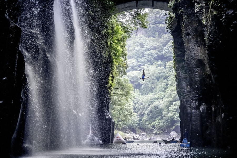 A high diver is seen in mid-jump, as they fall from a platform into a body of water between tall cliffs.