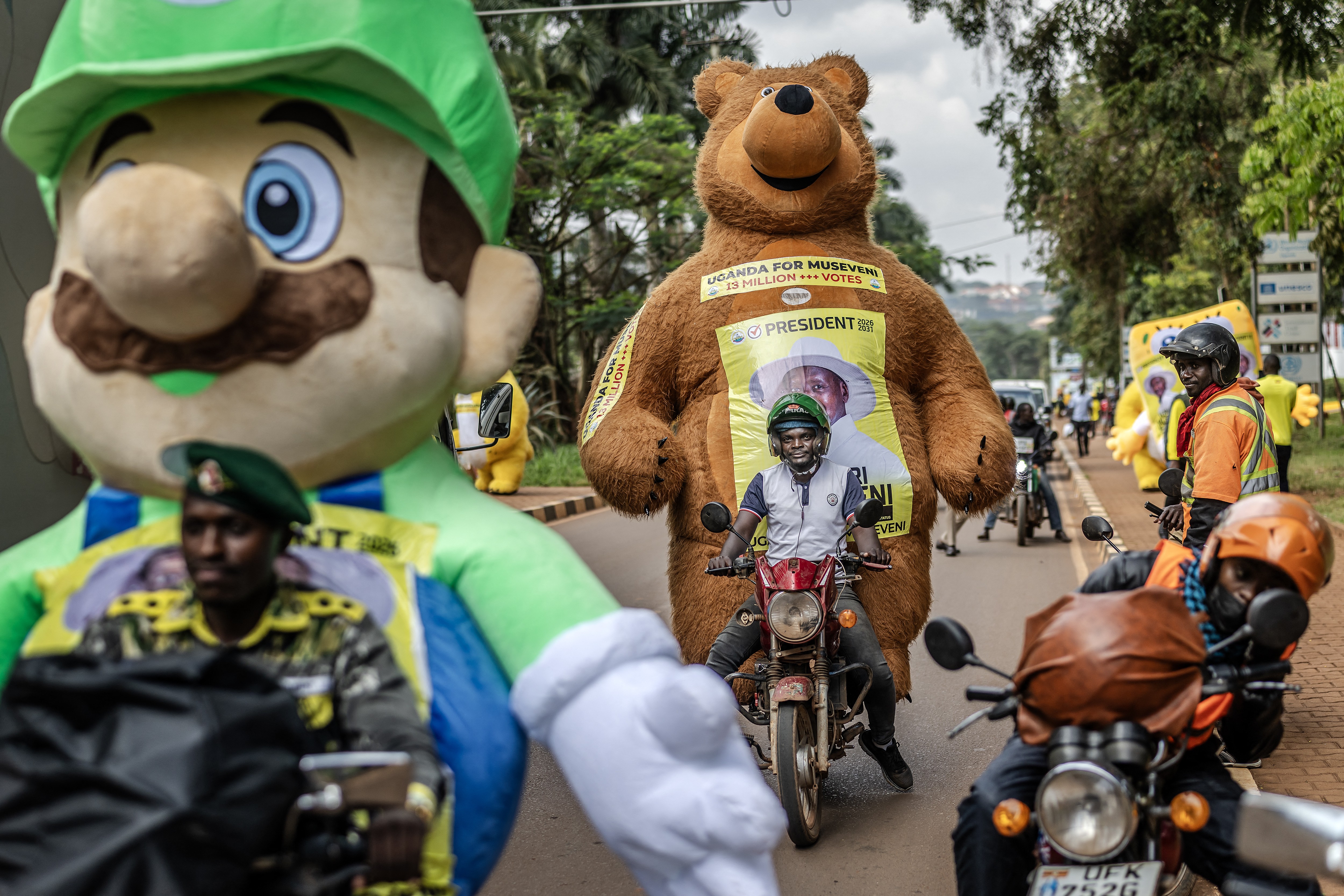 Two people on motorcycles carry large mascots as passengers, including an enormous stuffed bear, during a political campaign.