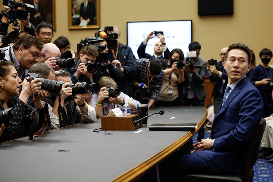 A person sits at a table for a hearing, photographed by more than a dozen members of the press.