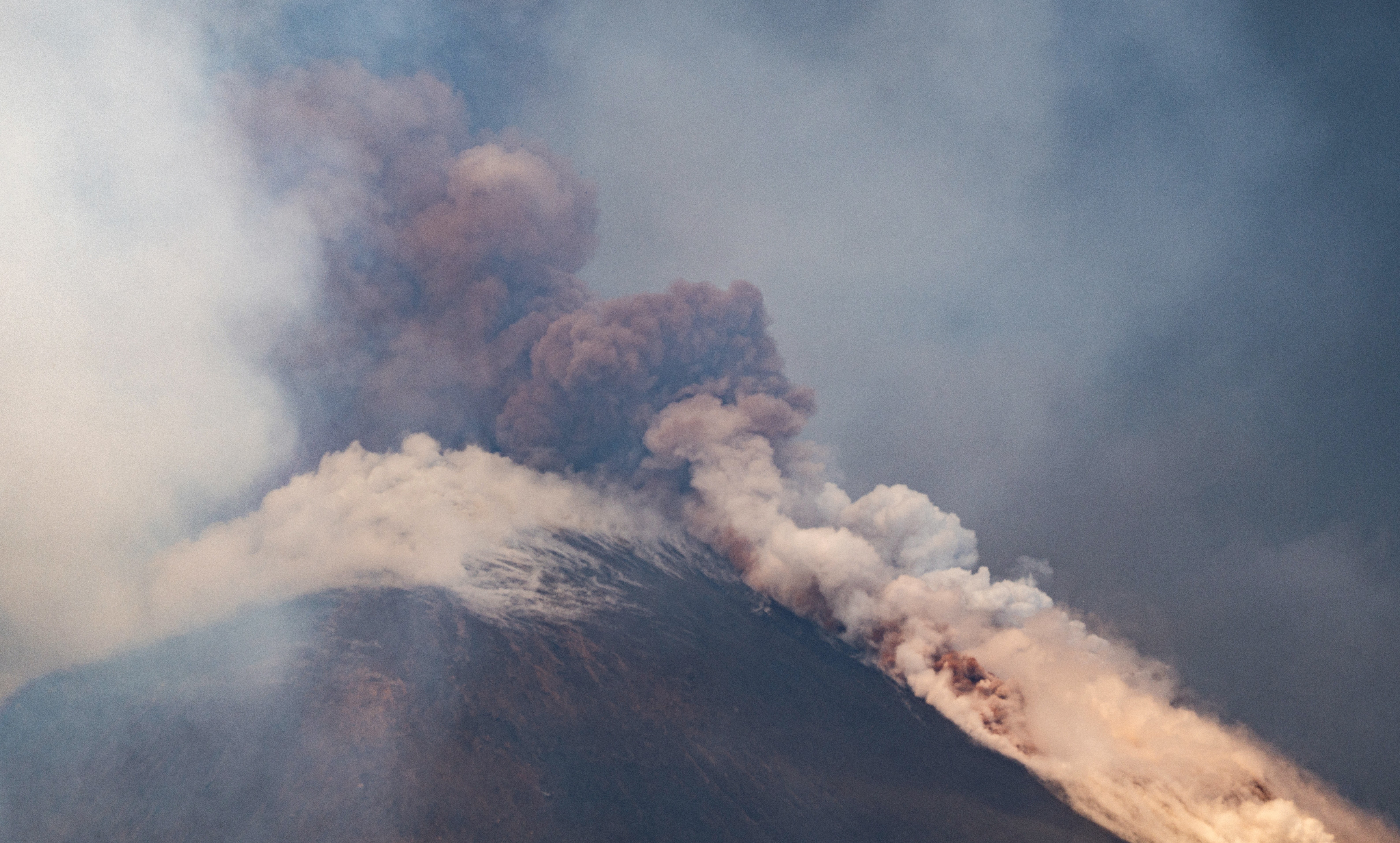 A plume of ash rises from a volcano, with steaming pumice surrounding its crater.