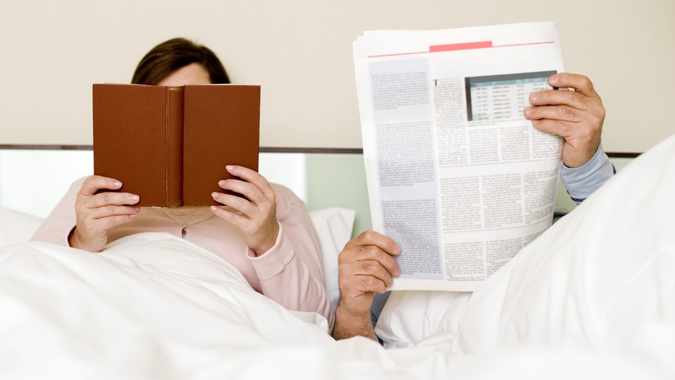 A couple sits in bed reading a newspaper and a book.