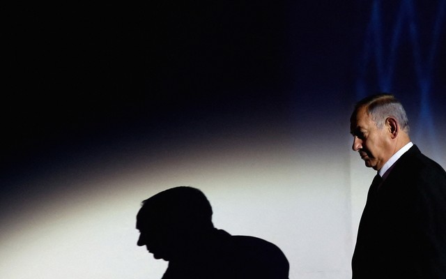 Photograph of Benjamin Netanyahu looking down with a spotlight shining on him and a shadow of himself projected on the wall behind him