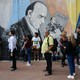 A loose queue of people in front of a wall featuring a mural of three men