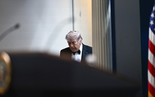 Trump in tuxedo entering White House briefing room; American flag visible on the side and podium in the foreground
