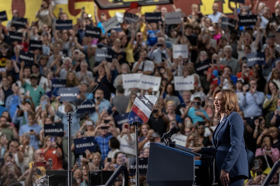 An enthusiastic crowd waves signs in a high-school gymnasium, as Vice President Kamala Harris stands at a podium.