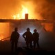 Firefighters stand in front of a burning house.