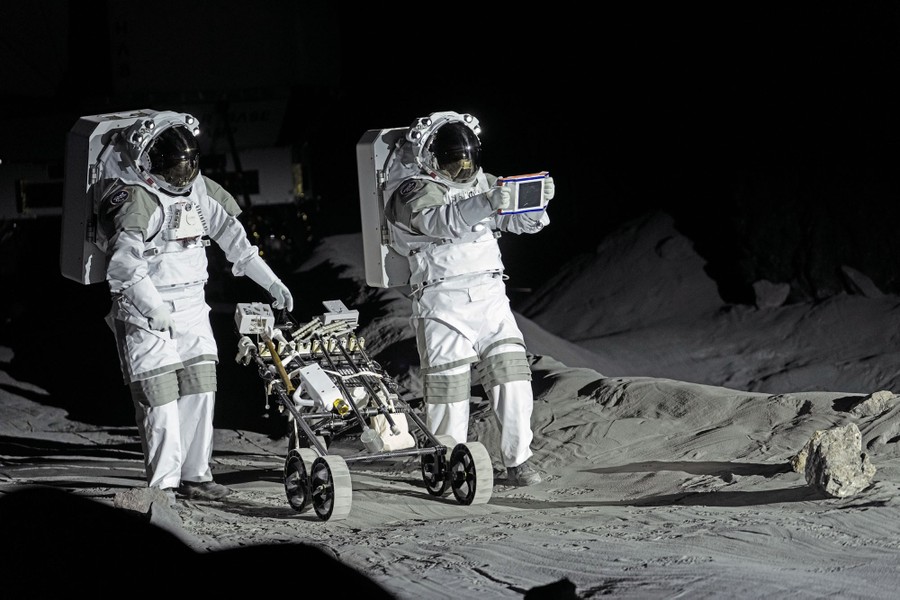 Two people wearing astronaut gear walk with a wheeled cart on a simulated lunar surface.