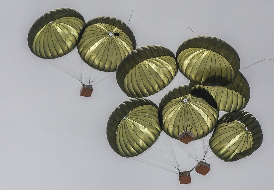 Several crates, suspended beneath pairs of parachutes, drop from the sky.