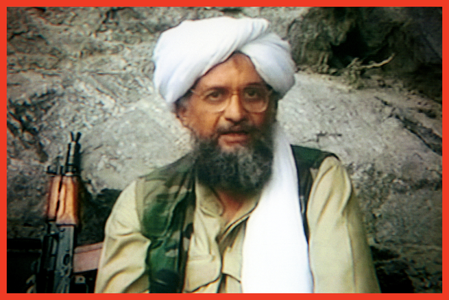 photo of bearded man in white head covering and tan jacket, wearing glasses and seated in front of rock face with rifle next to him