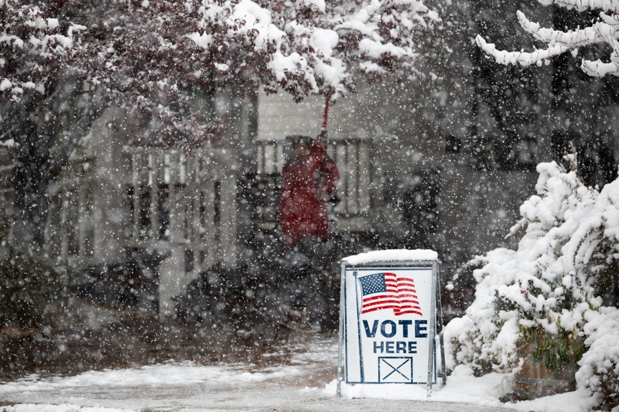 Snow falls outside a building with a "Vote Here" sign out front.