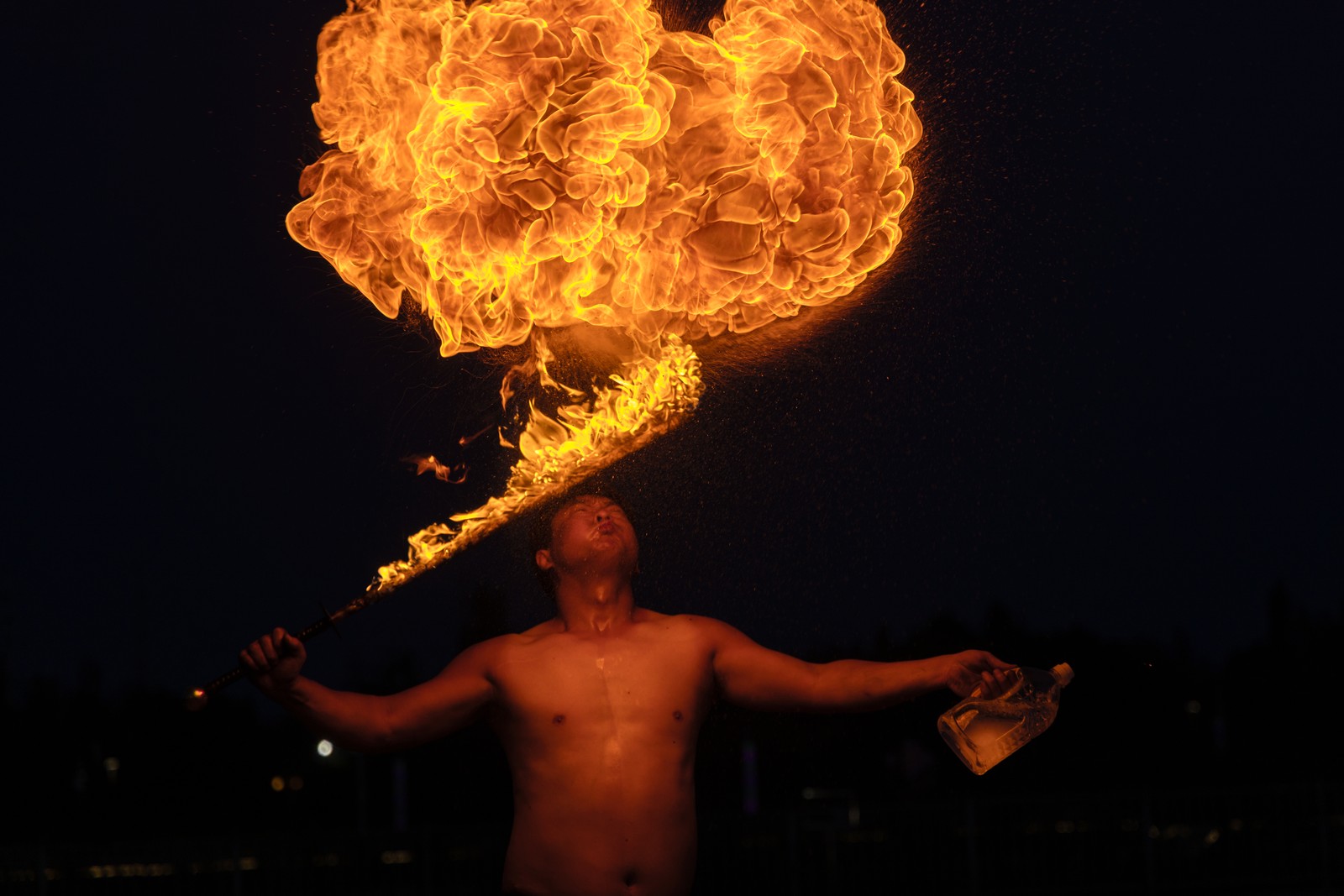 A performer holds a flaming sword while spitting a fireball.