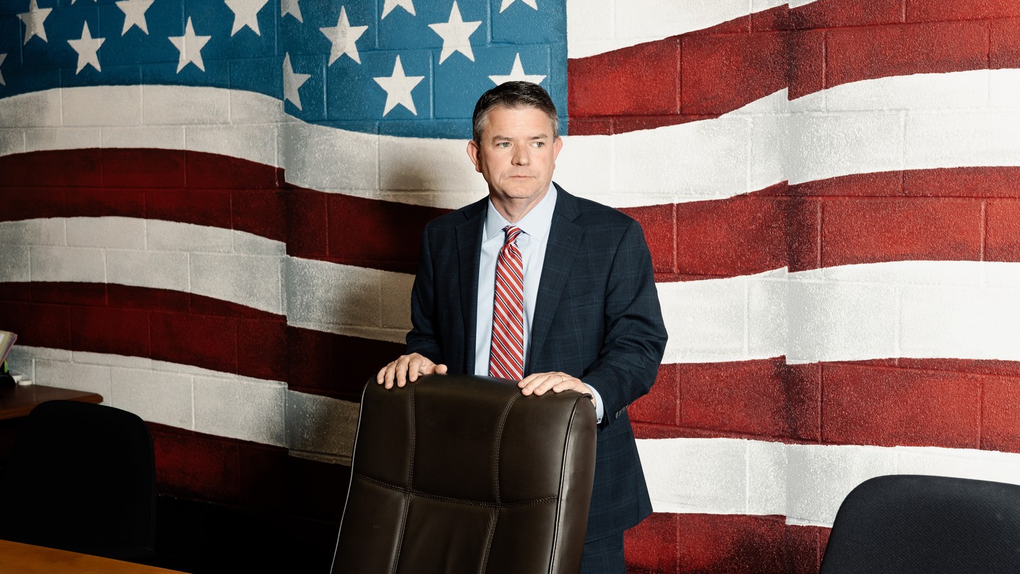Photo of a man standing behind a desk chair against a wall painted with a large mural of the American flag