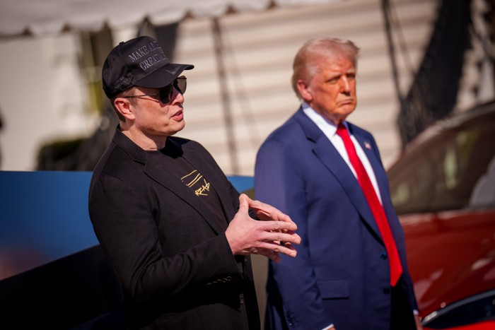 Elon Musk clasps his hands standing next to Donald Trump