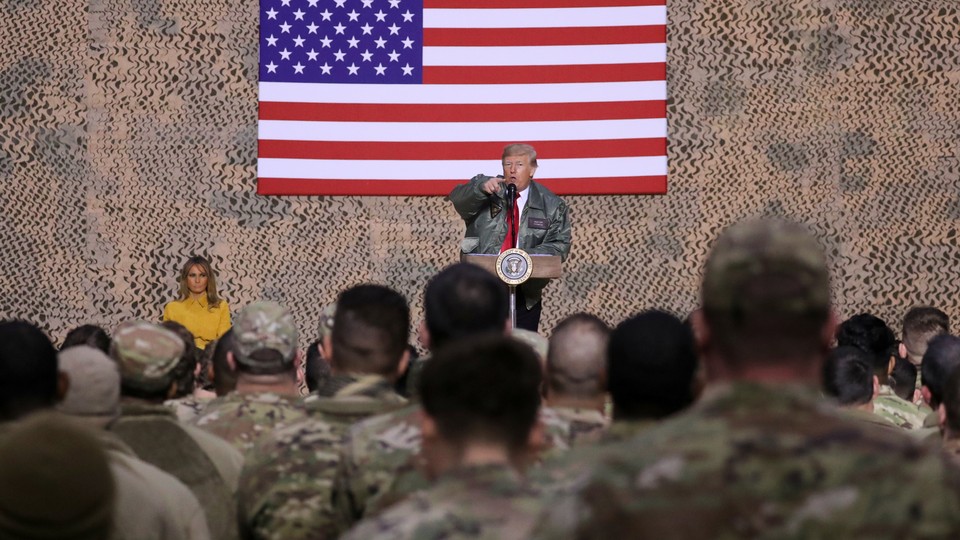 Donald Trump speaks to troops at al Asad Air Base with an American flag behind him and Melania Trump next to him.