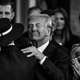 A black-and-white photo of Donald Trump facing the camera in a suit with a slight smile, while embracing Melania Trump, whose back is toward the camera. They are surrounded by people who are clapping and smiling.