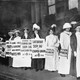 A black and white photo of women in dresses and feathered hats. They hold signs advertising an upcoming rally for women's right to vote.