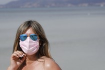 A woman on a beach in blue reflective sunglasses and a pink face mask