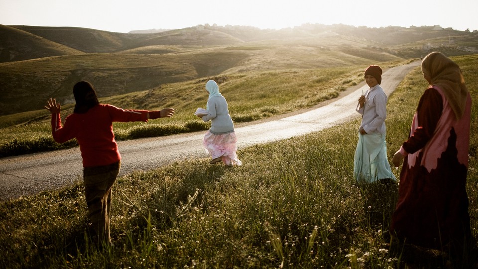 Girls walking along a road in Palestine, surrounded by rolling green hills, the sun beginning to set