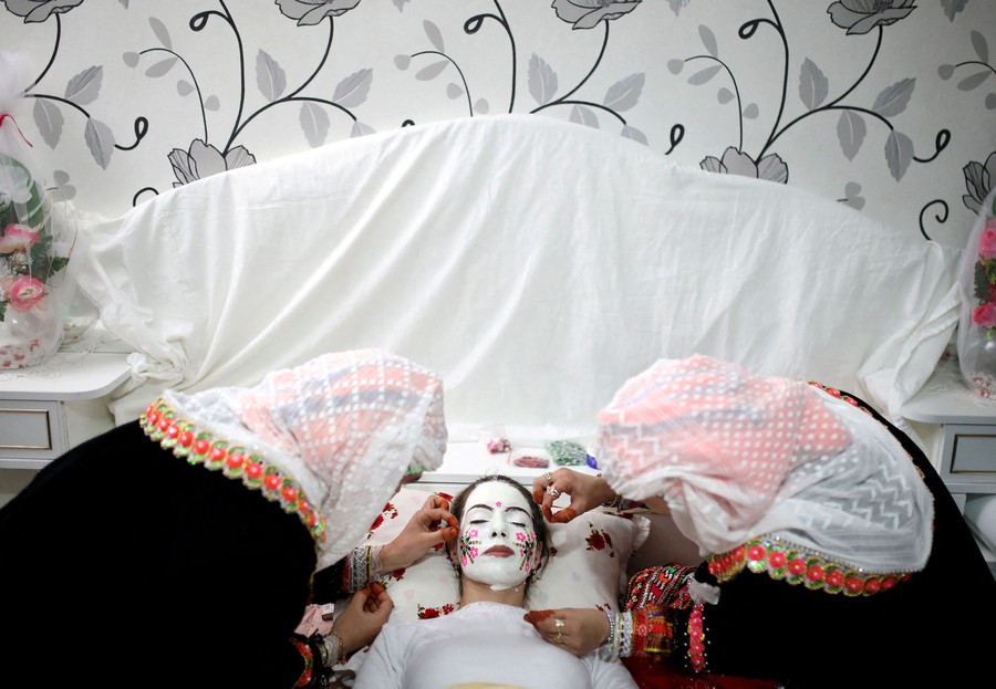 Two women lean over to apply white makeup and sequins to the face of a young woman lying on her back in a bed.