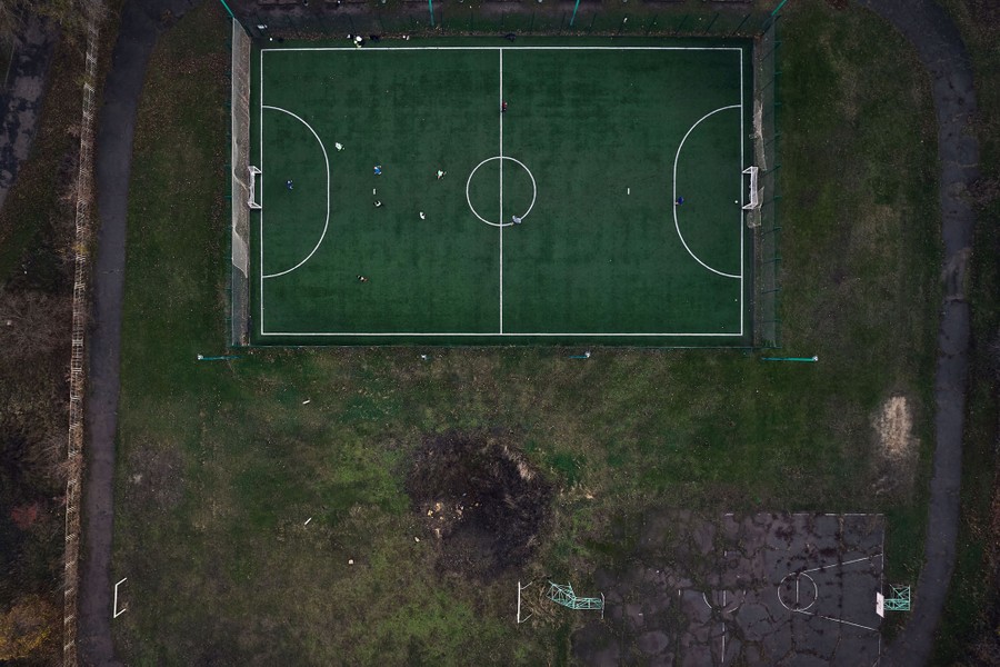 An aerial view of children playing soccer on a field beside a large bomb crater in Ukraine.