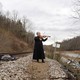 A person stands beside railroad tracks, playing a fiddle.