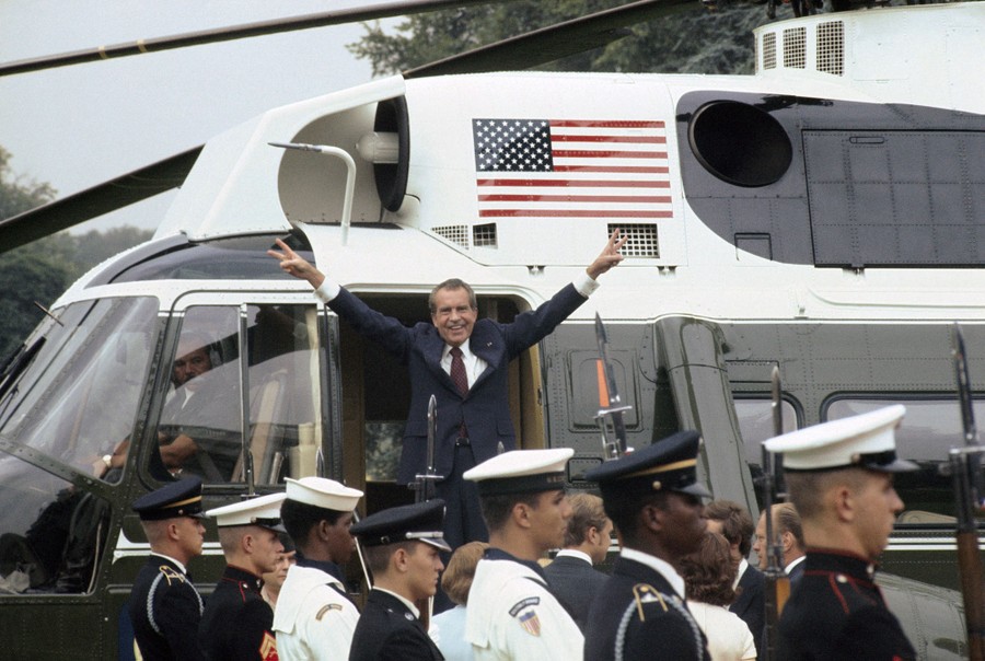 Former President Richard Nixon stands in the doorway of a helicopter, smiling and holding his arms up.