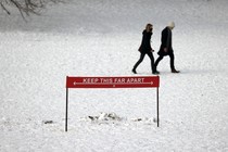 Two people walk together in a snowy NYC park behind a sign that says: "Keep this far apart."